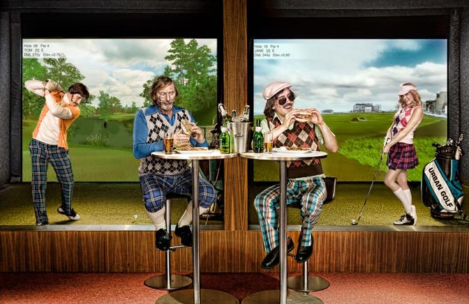 Two men and two women, dressed in golfing attire, are enjoying food and drinks in a golf-themed bar, surrounded by golfing equipment and screens displaying golfing simulations.
