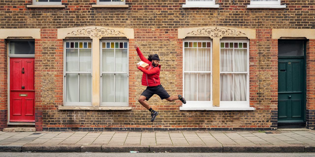 A man in a red jacket and black shorts jumps in front of a brick building with red and green doors.