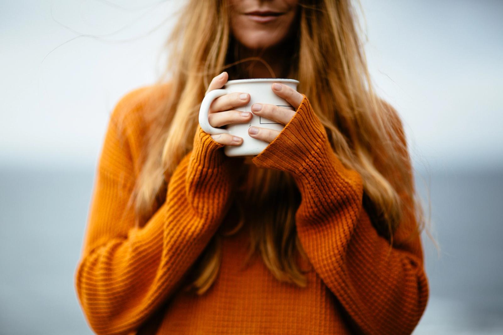 A woman holding a cup of coffee, wearing an orange sweater.