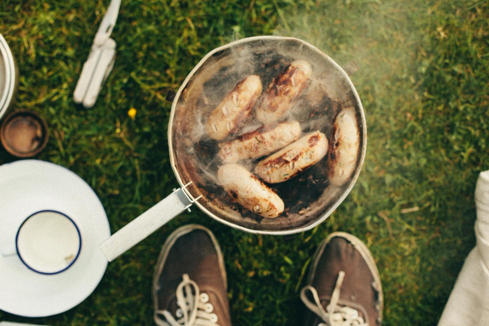 A pan of sausages being cooked on a grassy surface, with a pair of shoes and a plate of food in the background.