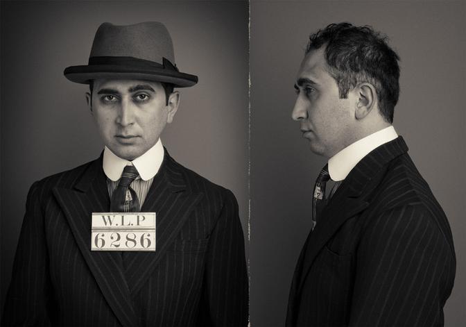 A man in a suit and fedora, with a fake mustache and a 'C' on his lapel, posing in a 1930s-style police lineup.