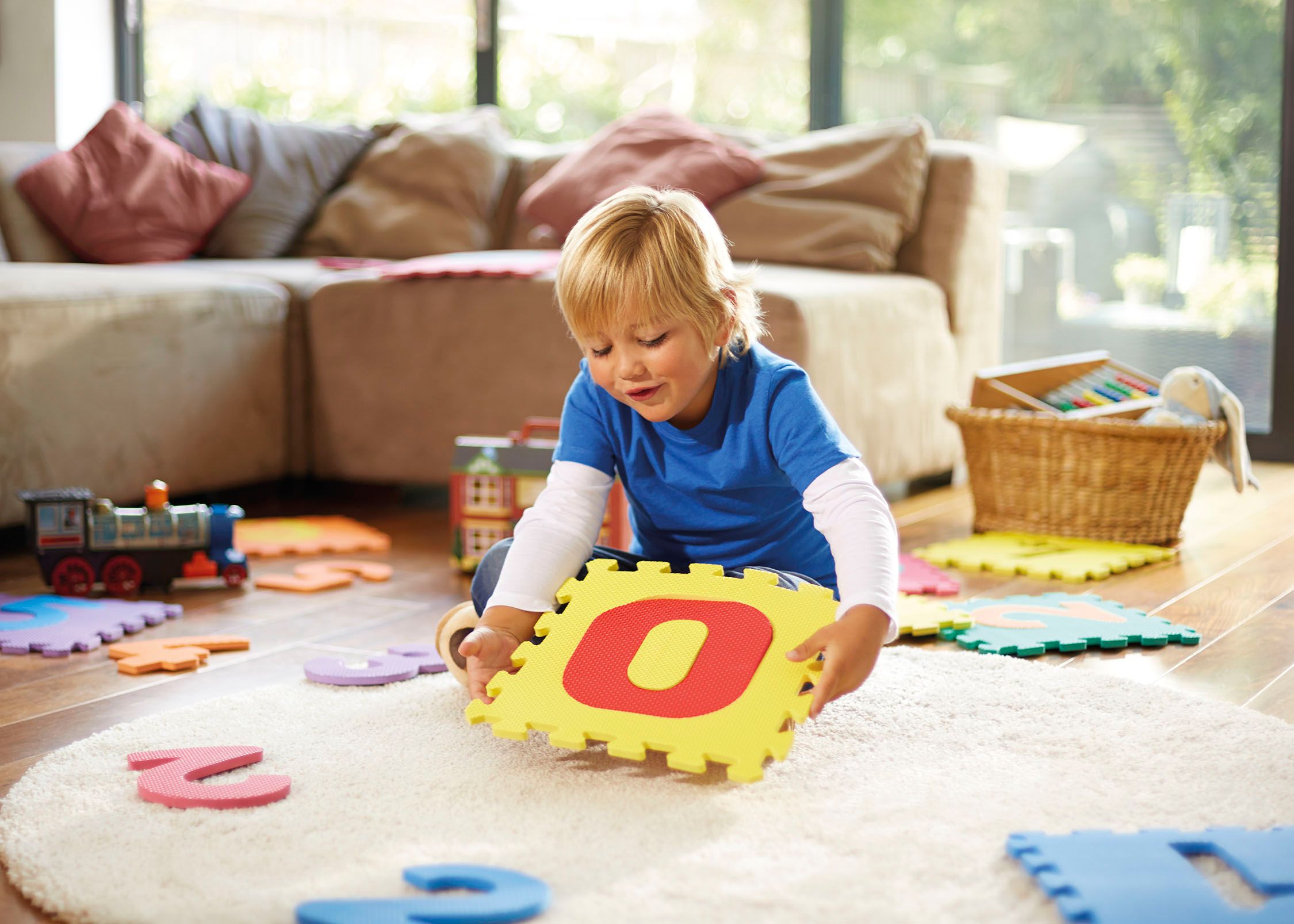 A young boy playing with a large, yellow, foam letter 'O' on a white rug in a living room with toys and a couch in the background.