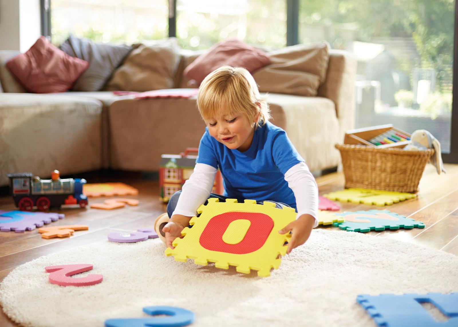 A young boy playing with a large, yellow, foam letter 'O' on a white rug in a living room with toys and a couch in the background.