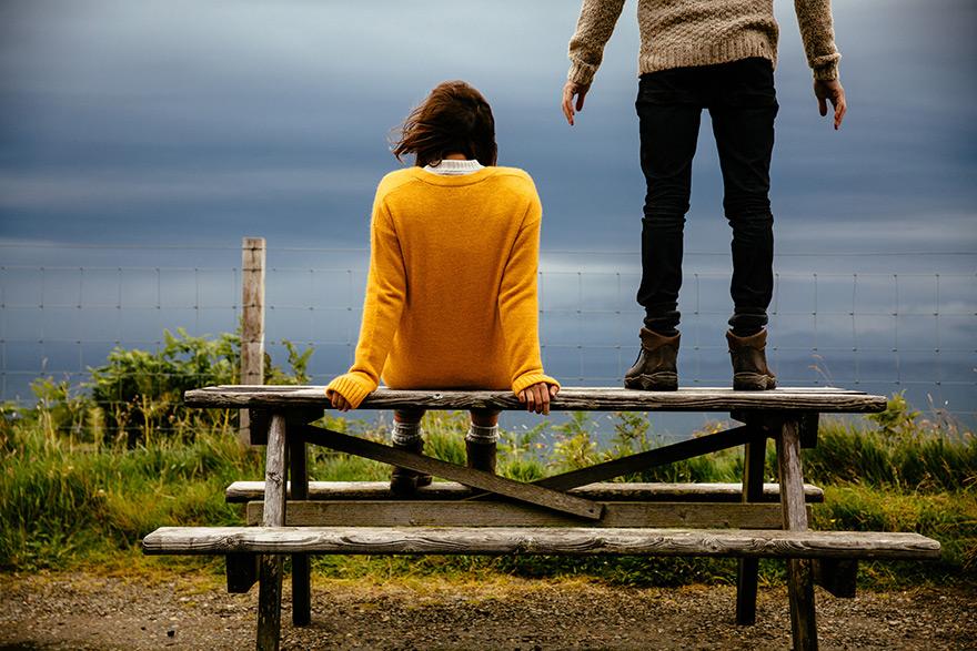 A woman in a yellow sweater sits on a wooden bench overlooking a vast, open landscape with a fence in the distance.