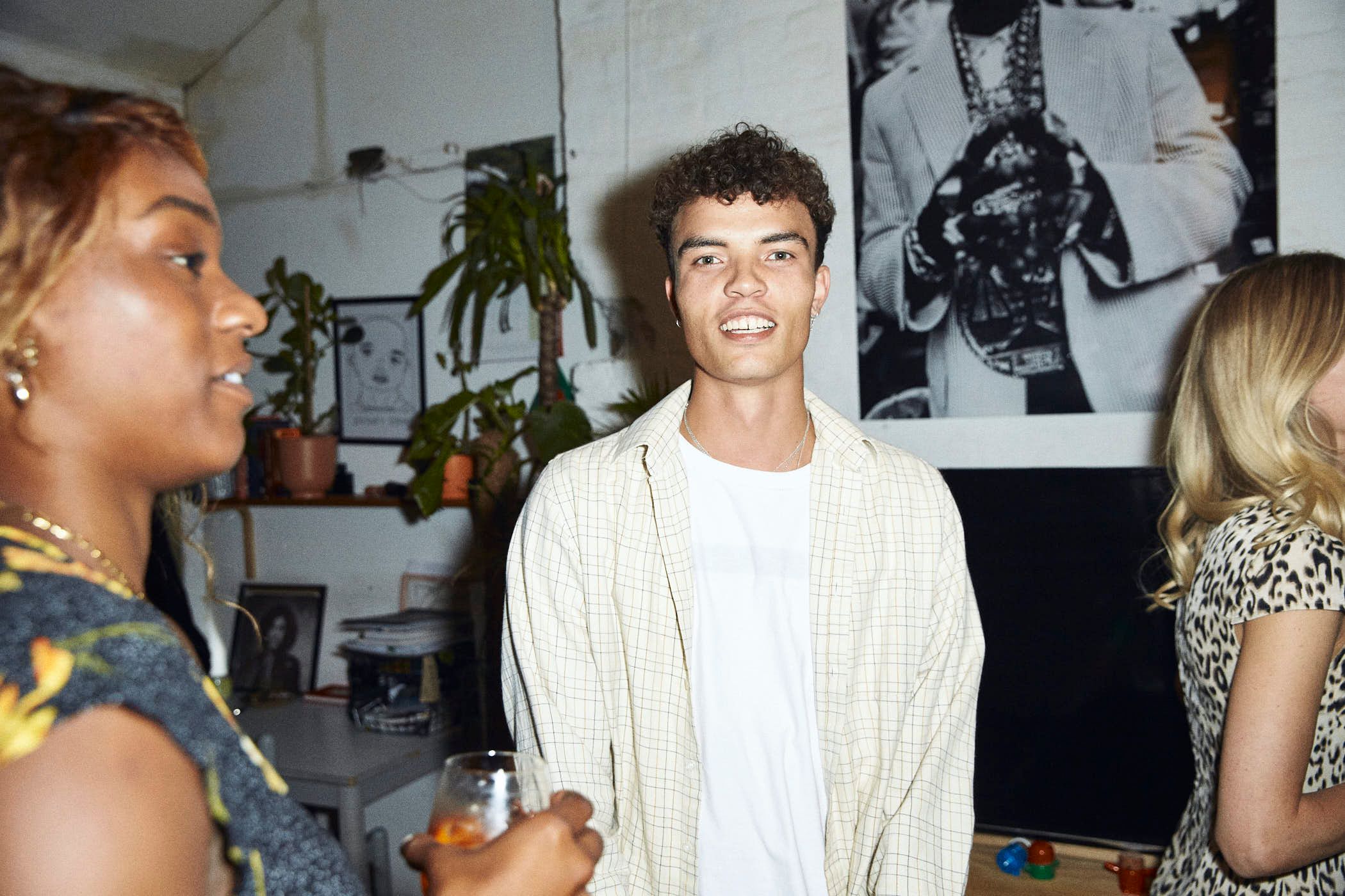 A group of people are gathered in a room with a mix of plants and artwork on the walls. The mood appears to be lively and social, with people smiling and holding drinks.