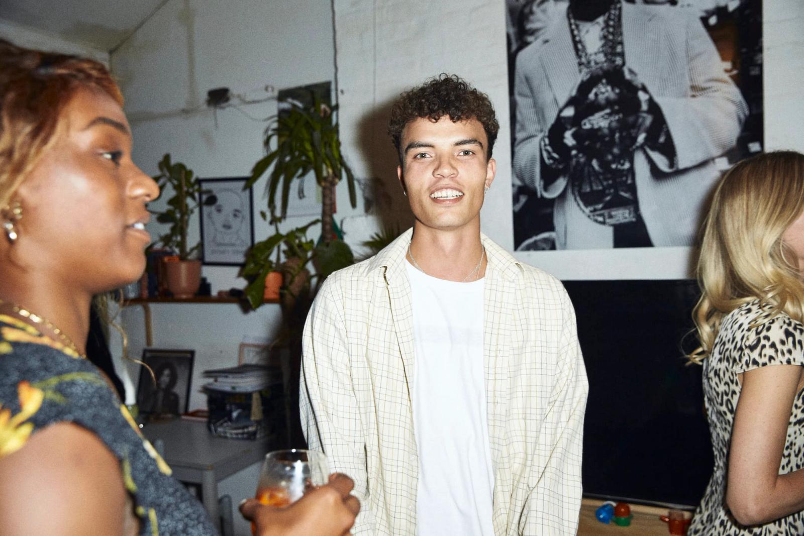 A group of people are gathered in a room with a mix of plants and artwork on the walls. The mood appears to be lively and social, with people smiling and holding drinks.