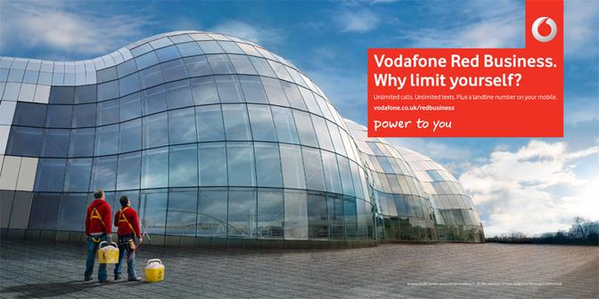 Two men in red Vodafone-branded jackets standing in front of a large, curved glass building, with a cityscape visible through the windows.