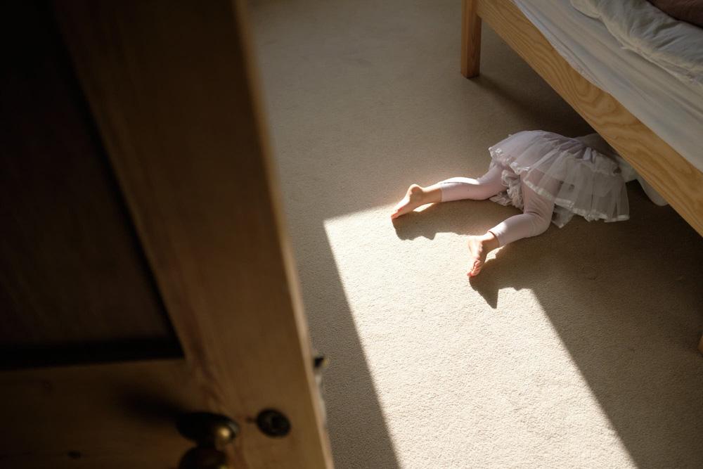 A child in a white tutu lies on the floor under a bed, her legs and feet visible in the sunlight. She appears to be playing or hiding.