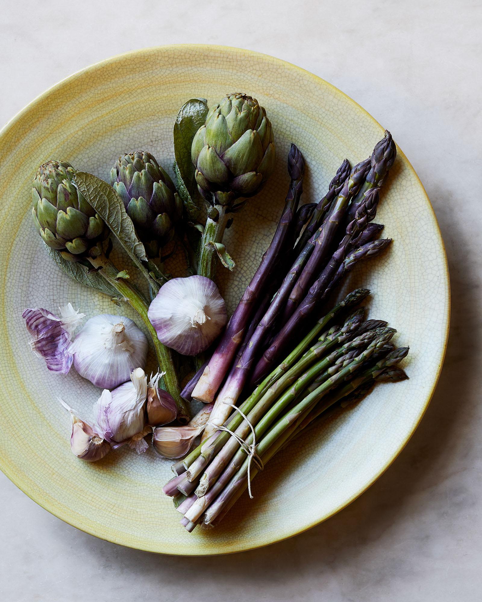 A selection of fresh vegetables, including artichokes, asparagus, and garlic, on a plate.