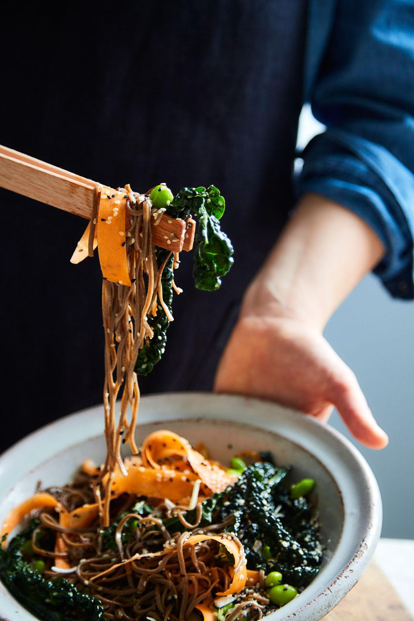 A person holds a large bowl of noodles with vegetables and chopsticks.