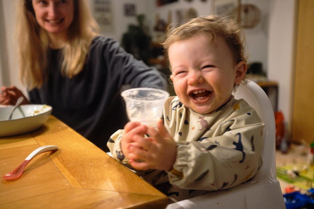 A young child in a high chair at a table, laughing and holding a cup. The child's mother is in the background, smiling and feeding the child.