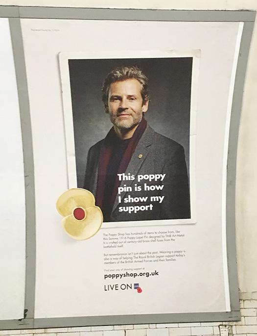A man wearing a grey suit, red scarf, and a gold poppy pin.
