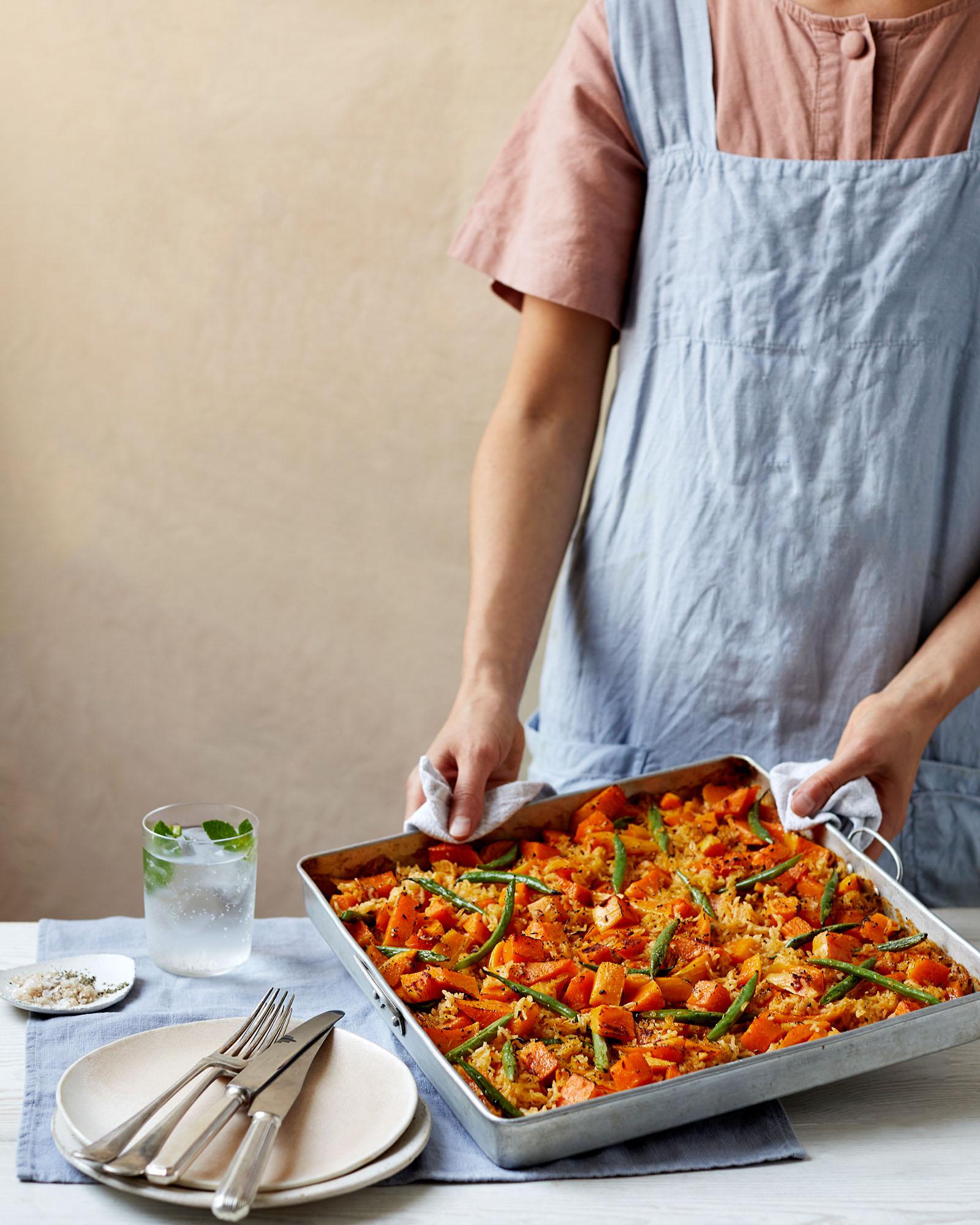A person in a blue apron is holding a large metal tray of roasted vegetables, including carrots, sweet potatoes, and green beans, on a white table.