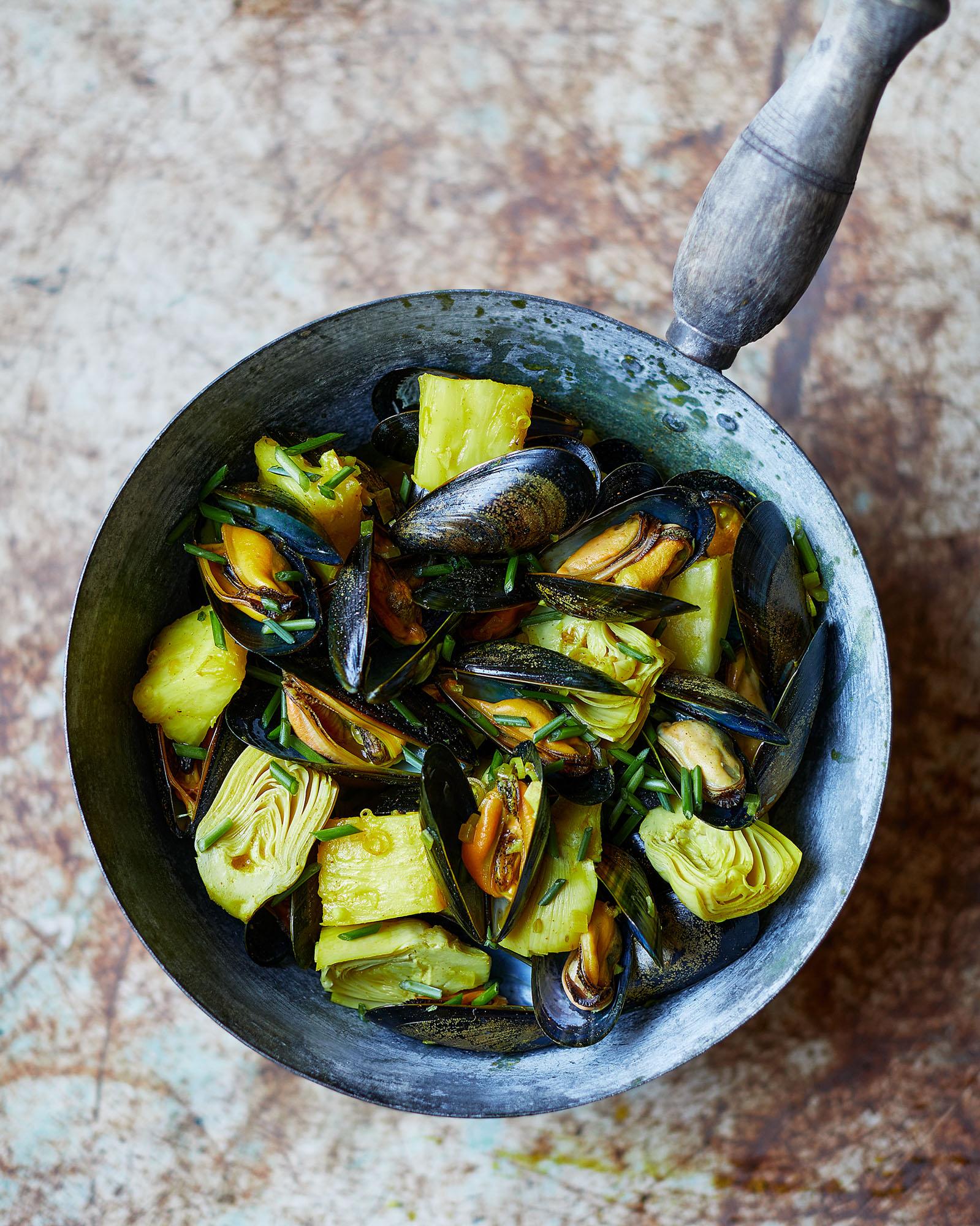 A large metal pan contains mussels, artichokes, and pineapple.