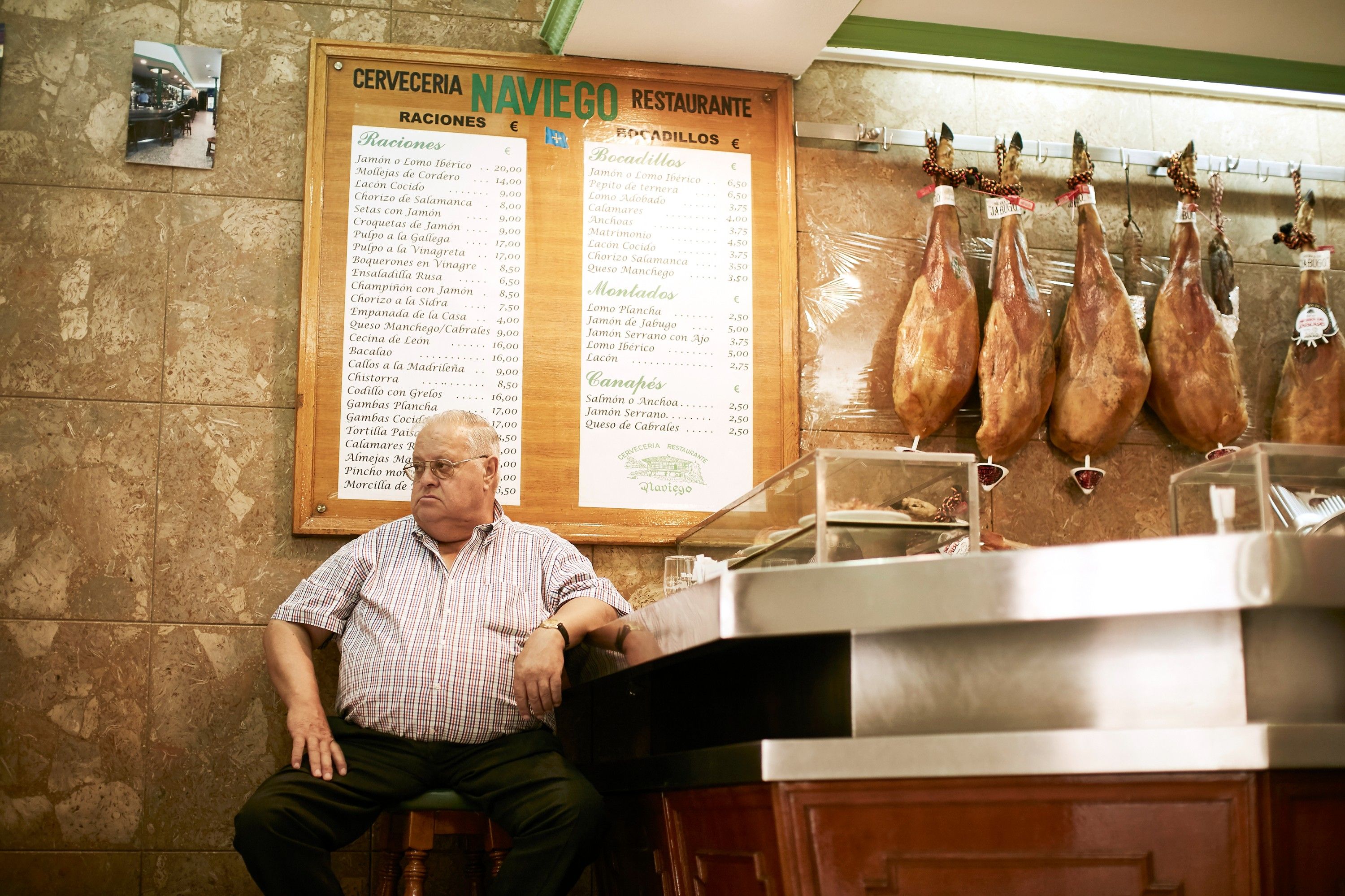 A man sits in a restaurant with a menu board displaying cured meats and Spanish menu items behind him.