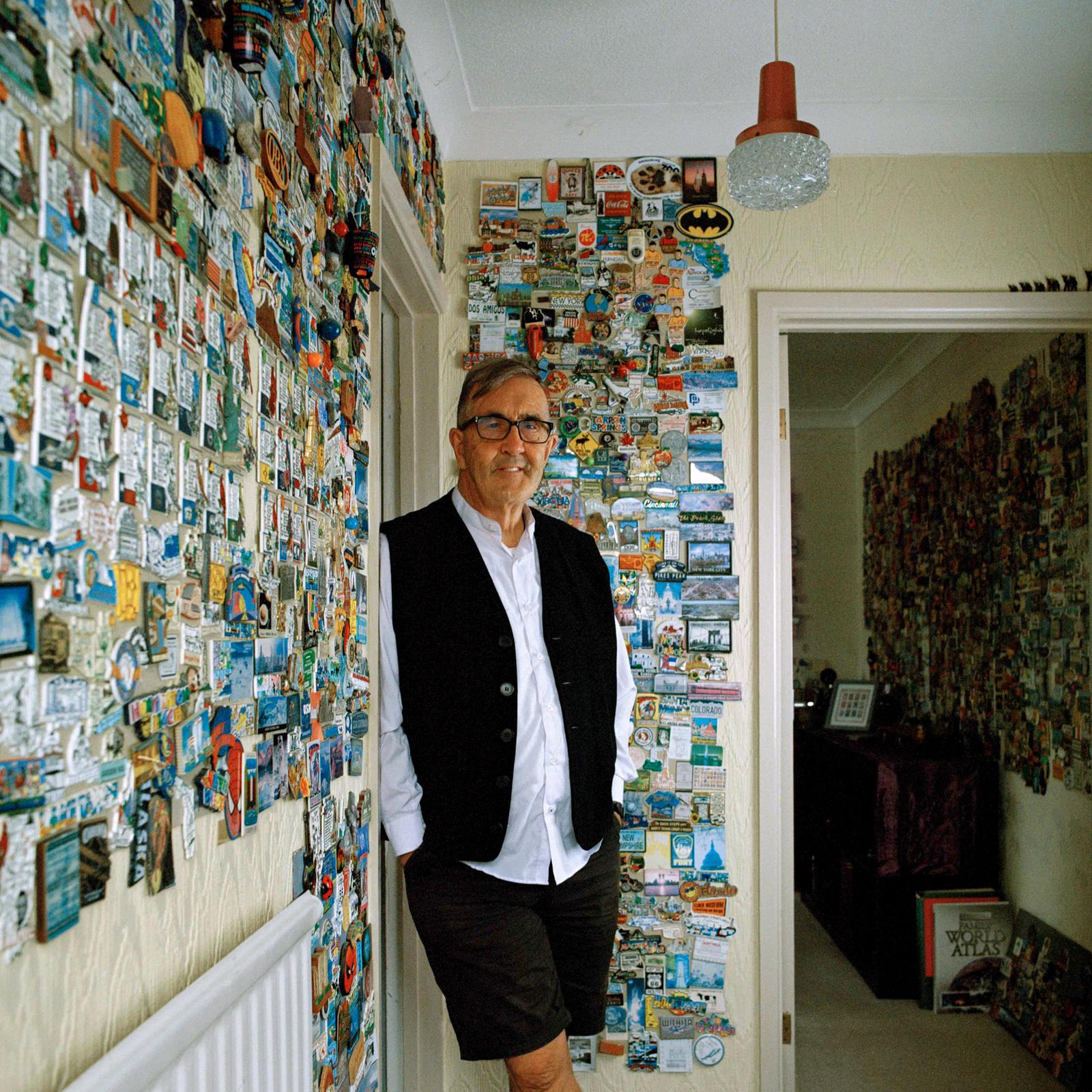 A man in a black vest and white shirt stands among a room filled with postcards and stickers.