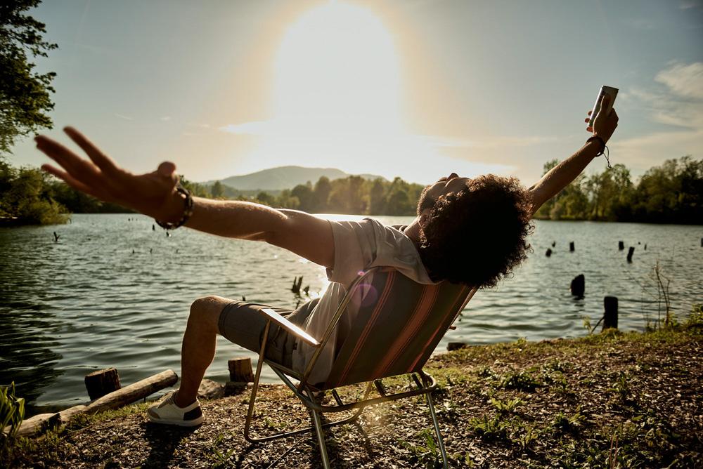A man sits in a chair by a lake, arms outstretched, holding a drink, during a sunset.
