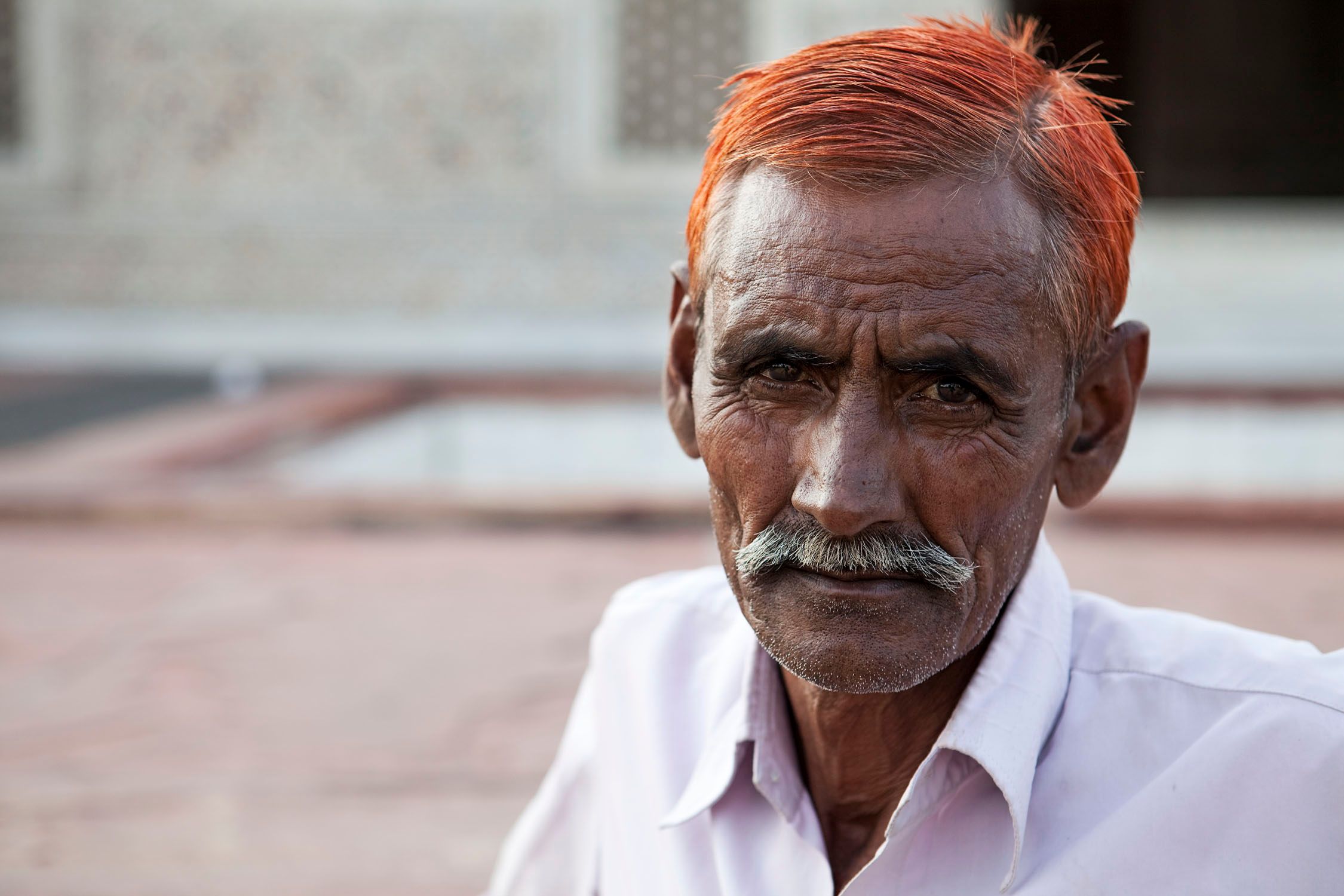 A man with a white shirt and long white mustache stands in front of a blurred stone building.