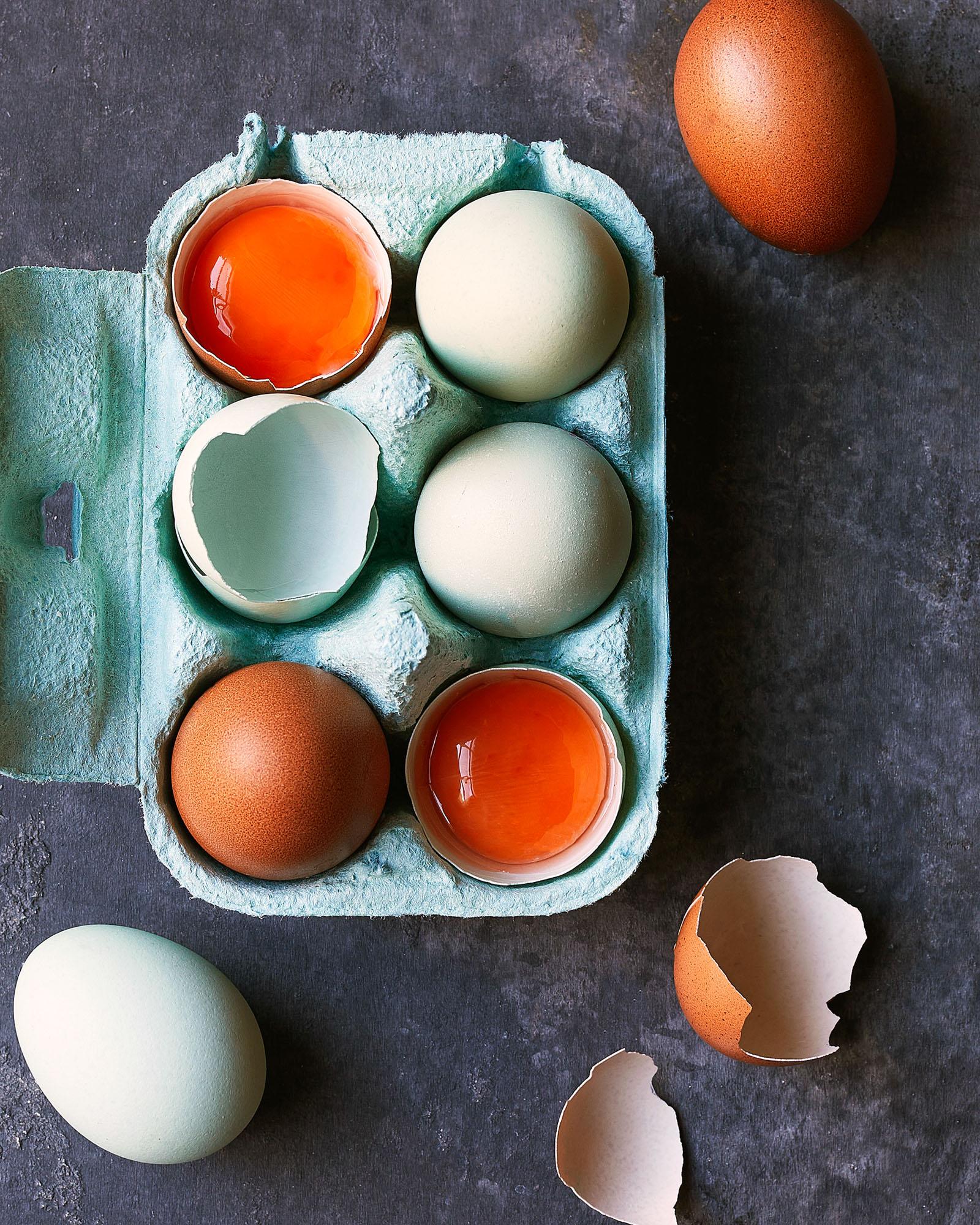 A selection of six eggs, three brown and three blue, in a carton with the carton's top flaps open, showing the eggs' contents are empty and the eggshells are broken.