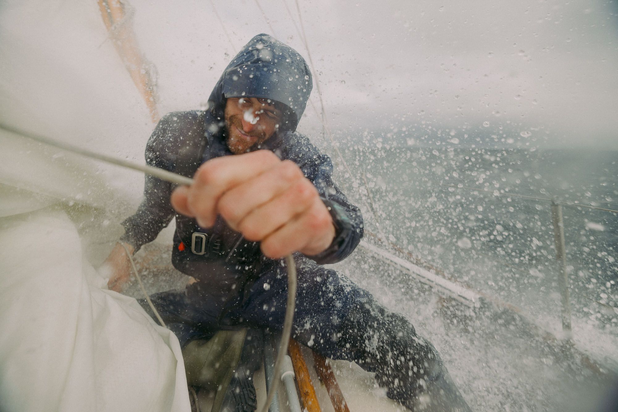A man in a wetsuit is holding onto a rope on a boat in rough seas, with water splashing everywhere.