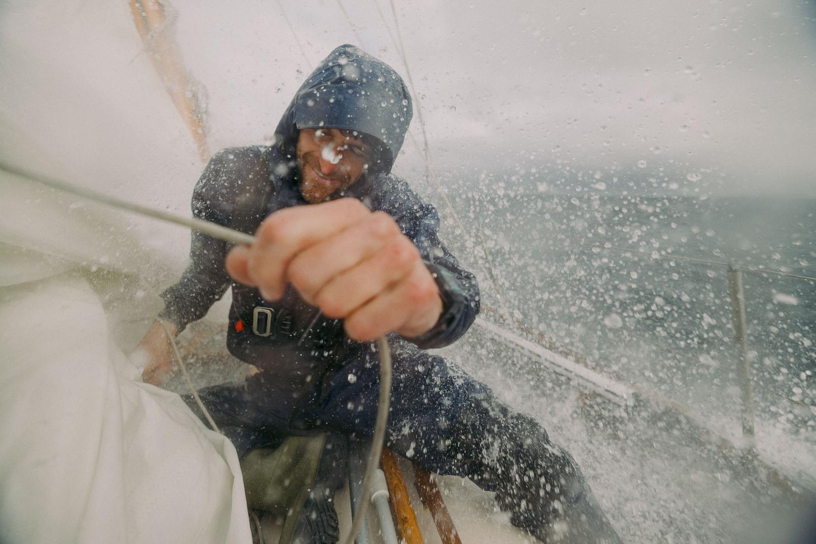 A man in a wetsuit is holding onto a rope on a boat in rough seas, with water splashing everywhere.