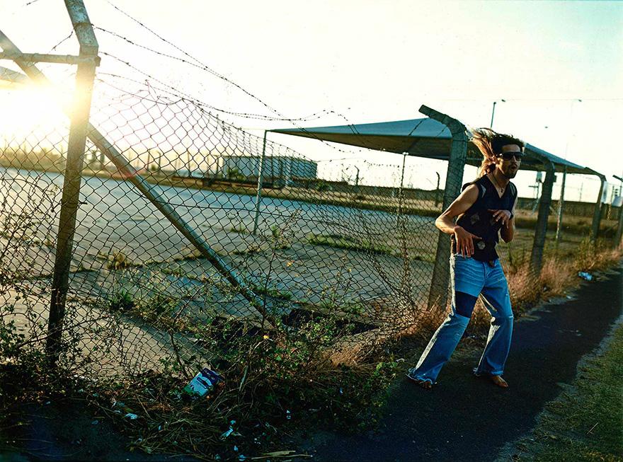 A man in a black tank top and blue jeans is running in an abandoned, fenced-in area with a chain-link fence and a large, empty parking lot in the background.
