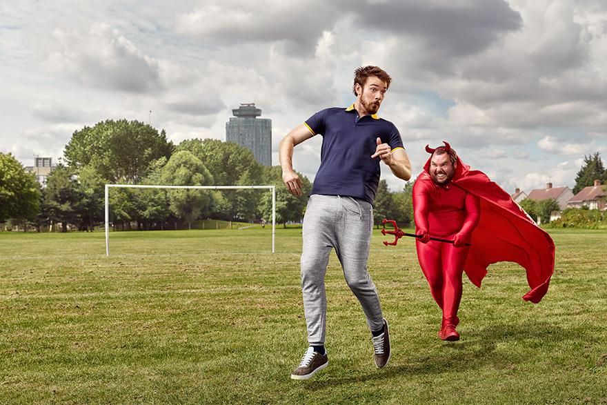 A man in a football jersey and grey sweatpants is running on a grassy field with a man in a red devil costume and a pitchfork running behind him.