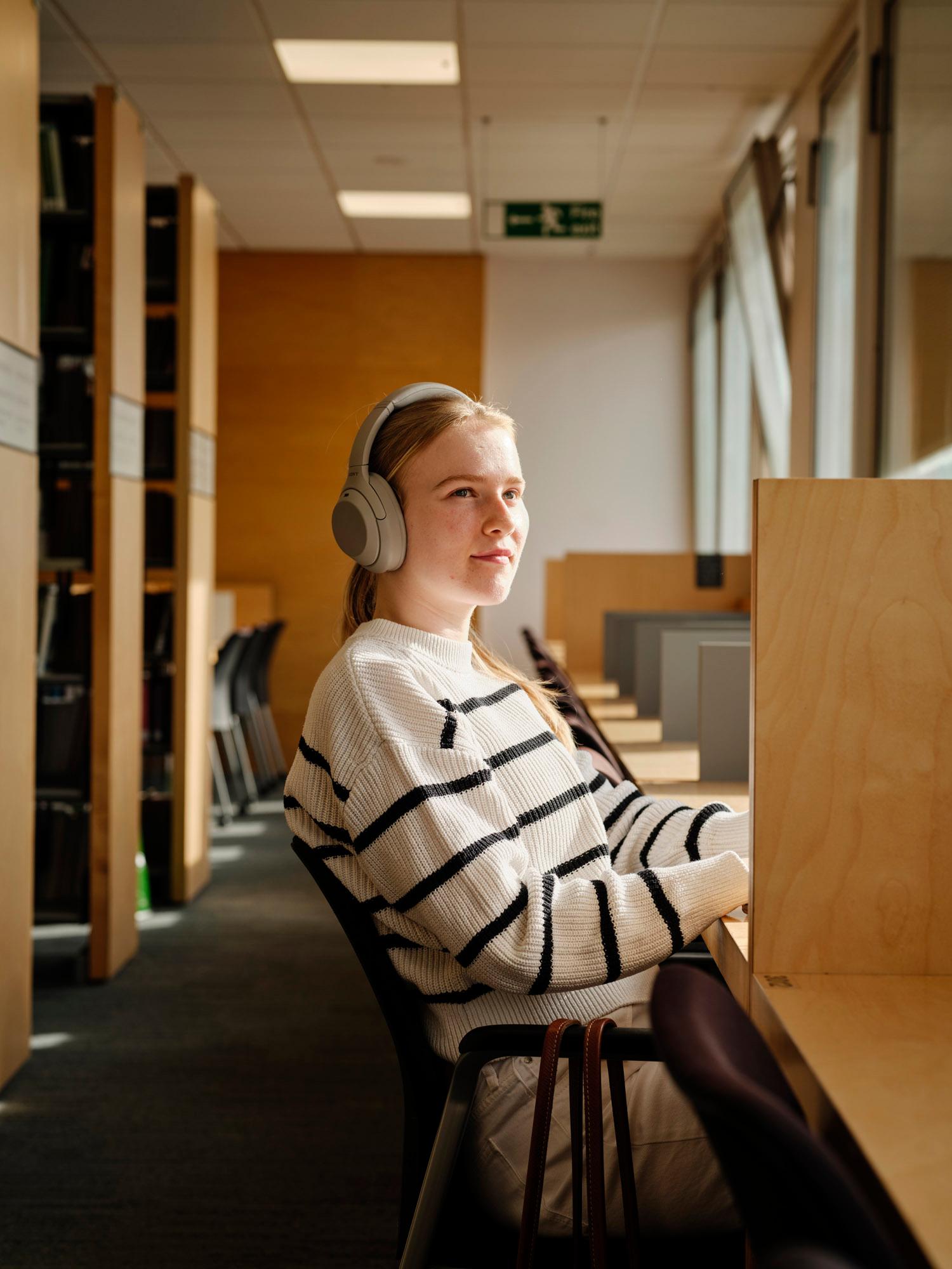 A young woman with long blonde hair and a white sweater with black stripes sitting in a library or study room, wearing large grey headphones.