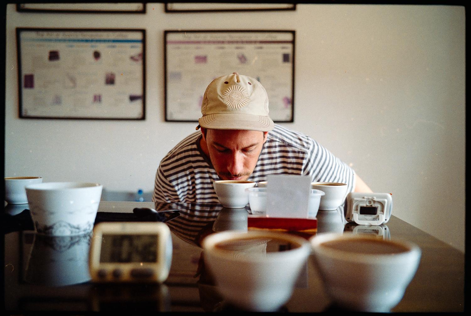 A man in a striped shirt and a baseball cap is leaning over a table, inspecting a bowl of food.