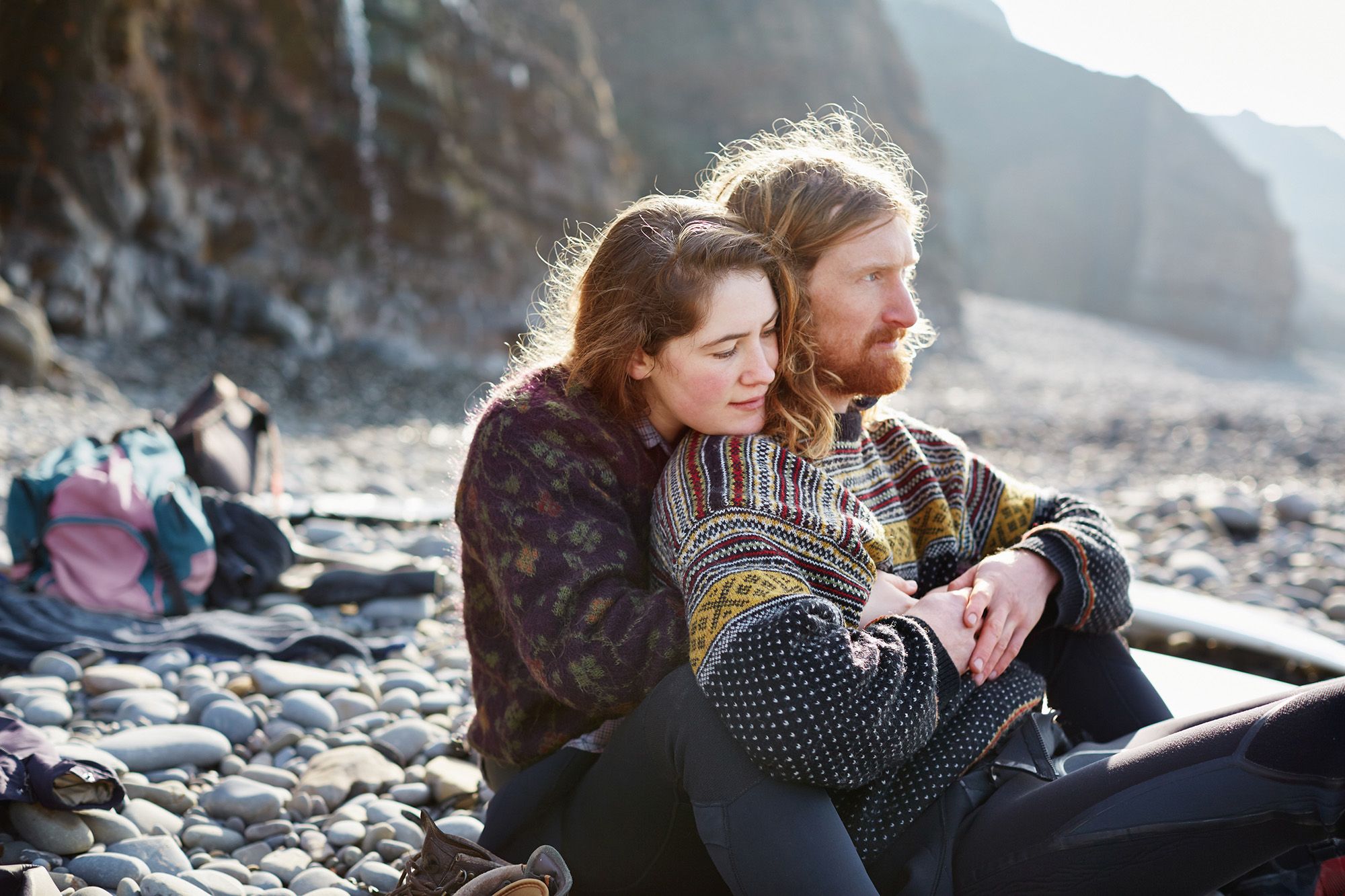 A woman and a man sitting on a rocky beach, embracing and looking out at the sea.