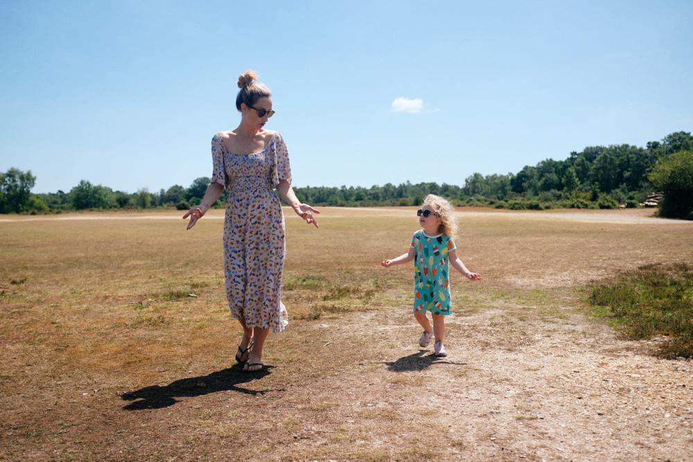 A young mother and child walk hand in hand through a vast, dry field on a sunny day, both wearing long dresses and sunglasses.