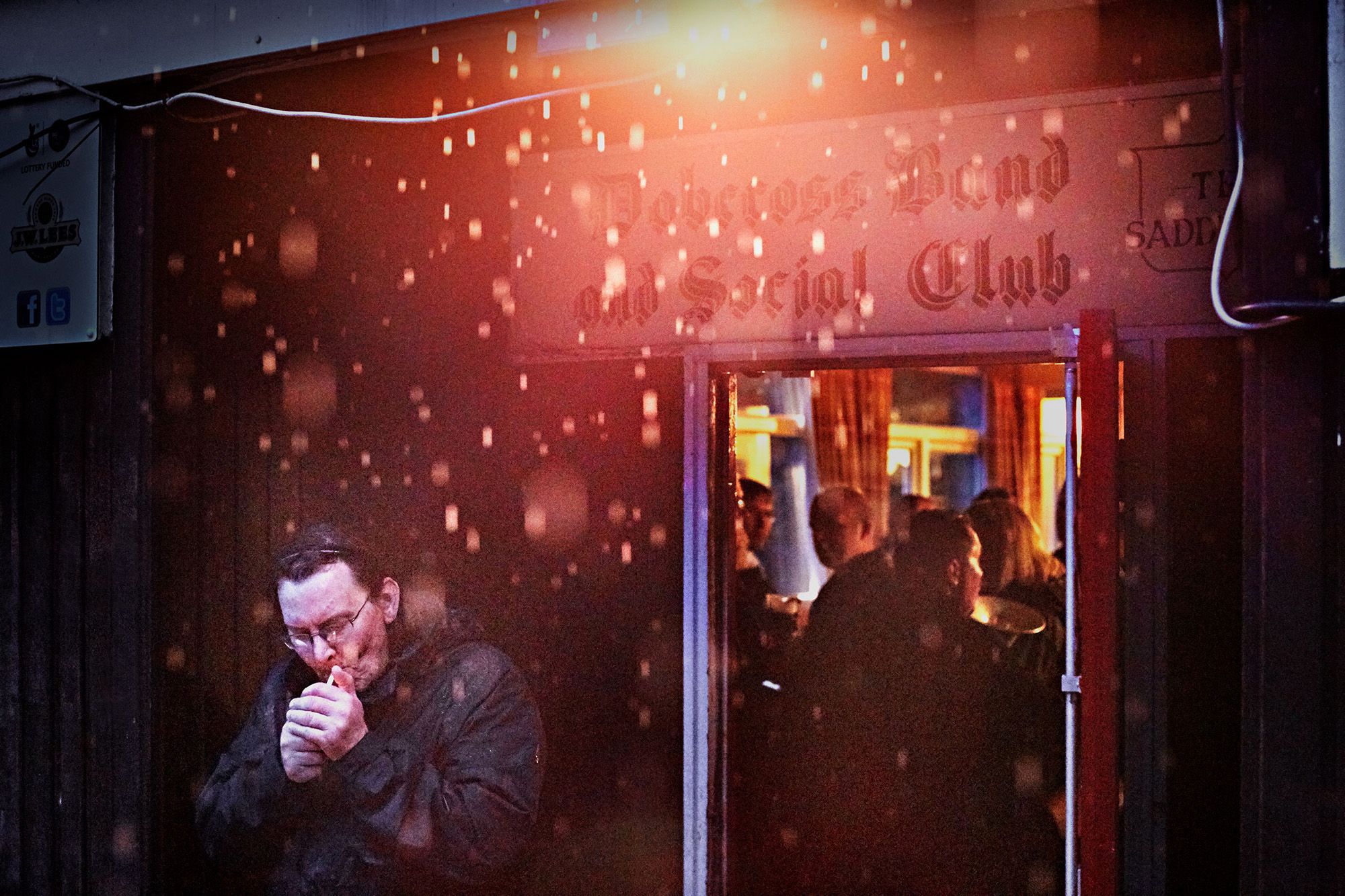 A man in a black jacket and glasses is standing in front of a nightclub entrance, smoking a cigarette.