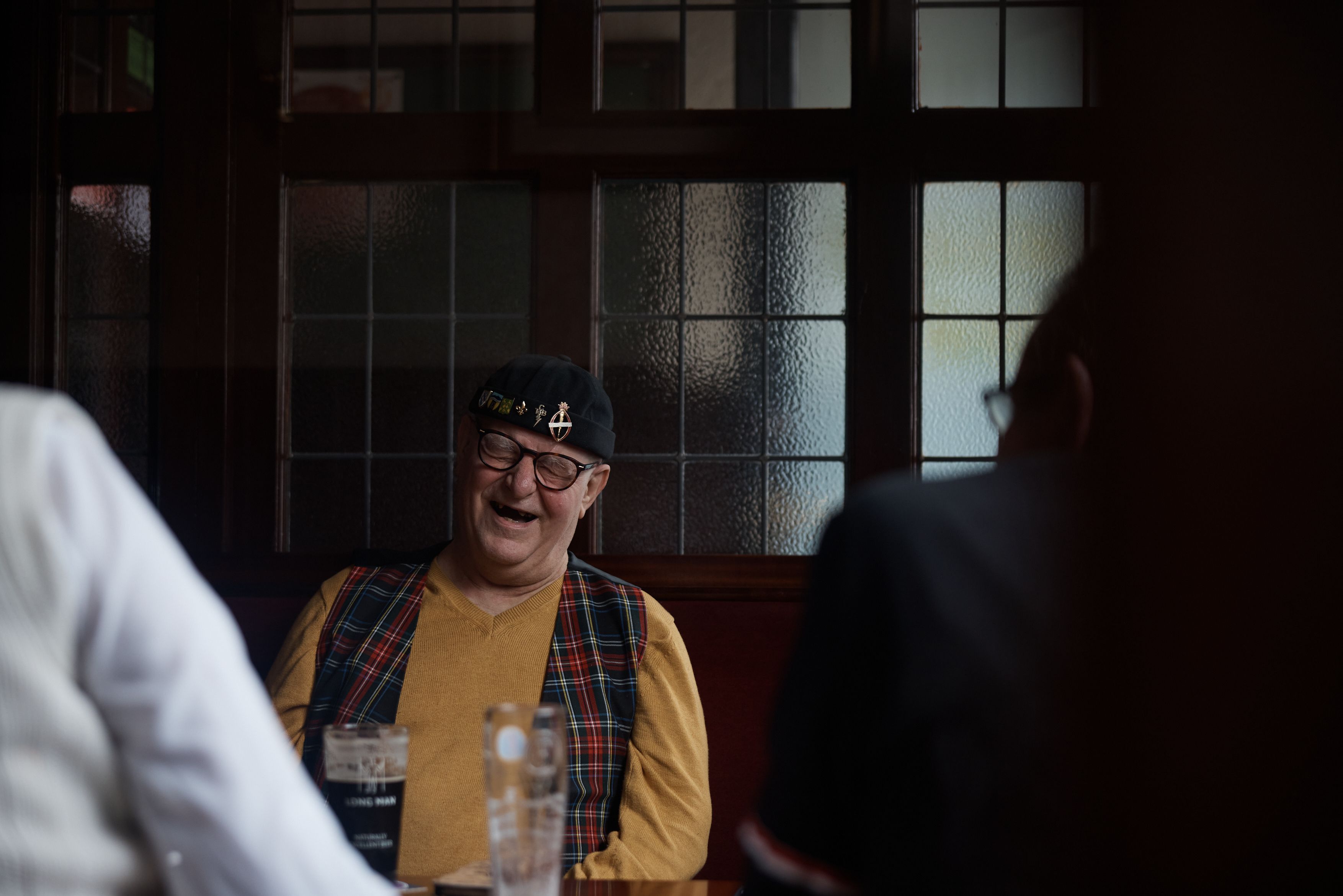 A man in a yellow shirt with a plaid vest and a black hat with a silver pin on it, sitting in a dimly lit pub, surrounded by people and glasses of beer.