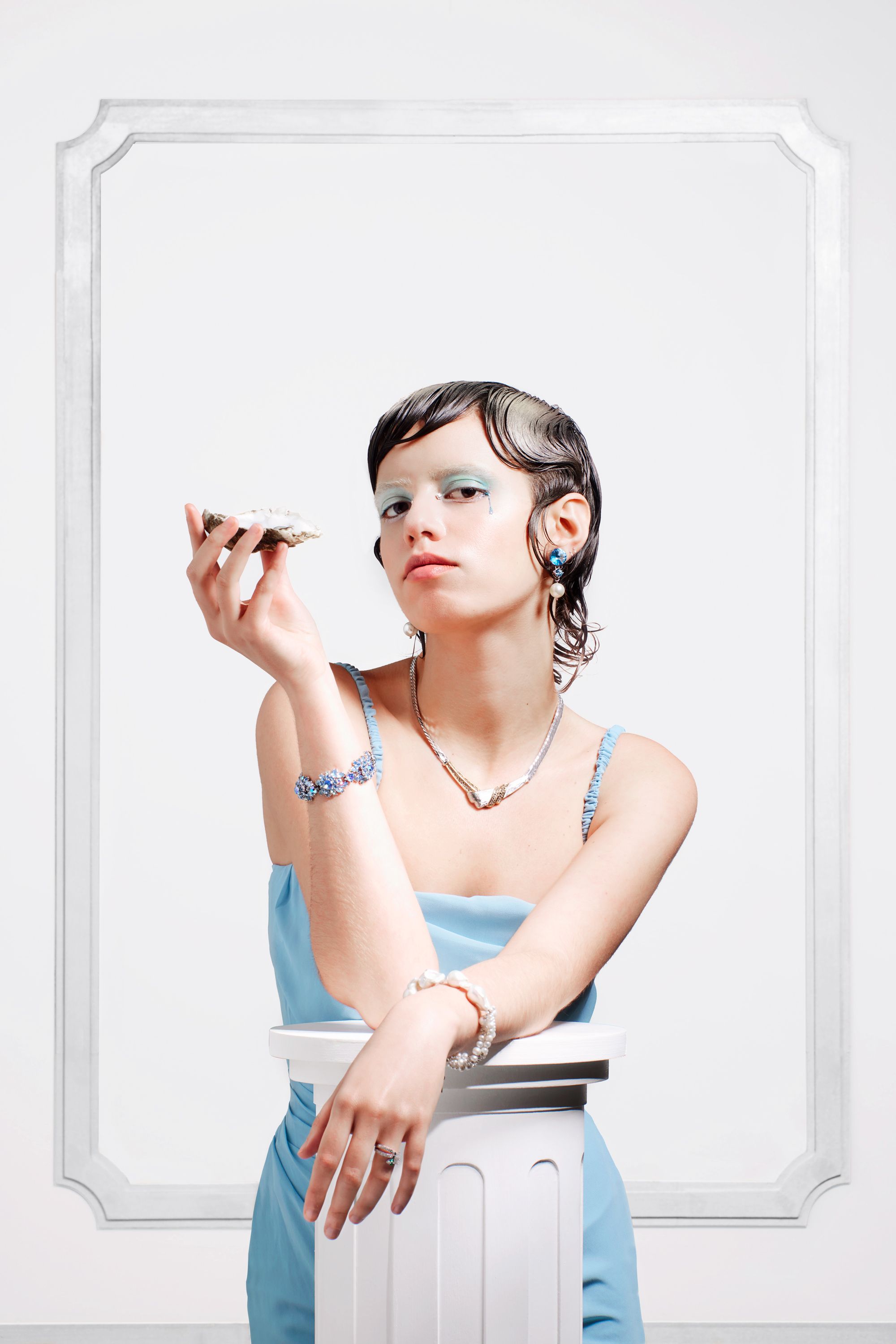 A woman in a blue dress holds a seashell to her ear, looking wistful.