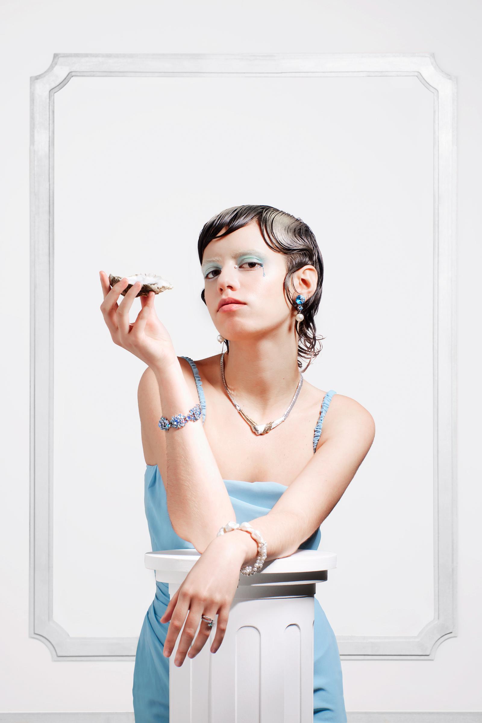 A woman in a blue dress holds a seashell to her ear, looking wistful.