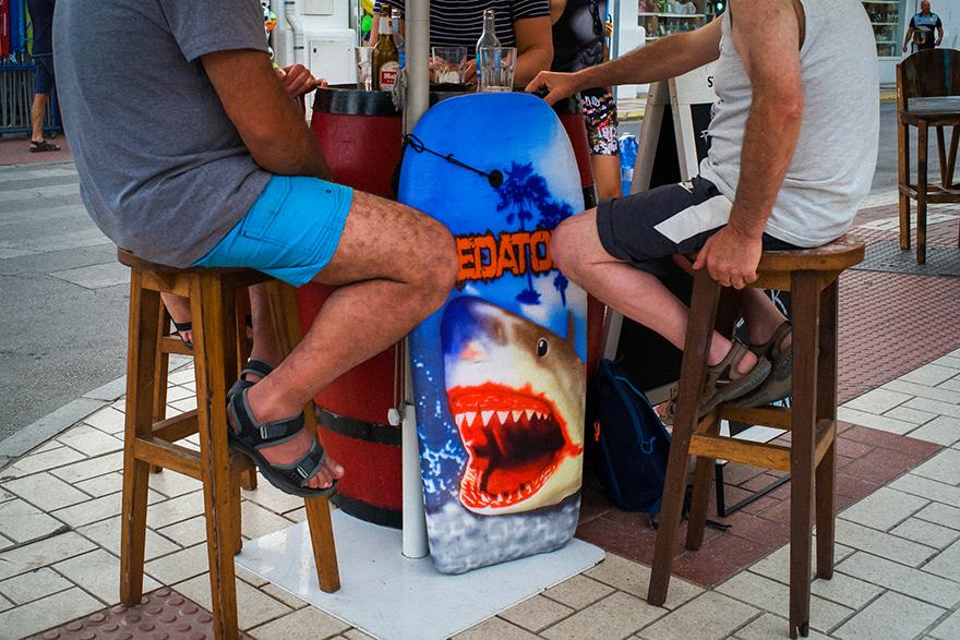 Two men sit on stools at a bar, one in a grey t-shirt and blue shorts, the other in a white t-shirt and black shorts; both have feet on a table with a surfboard featuring a shark.