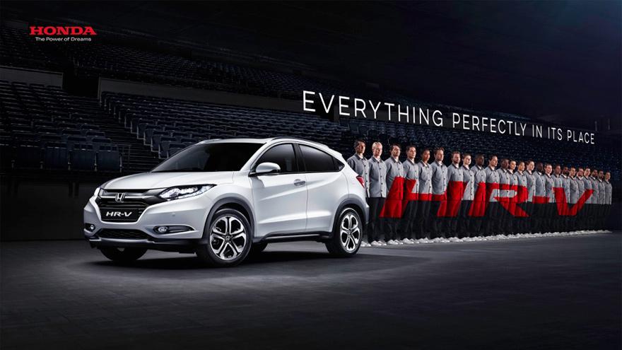 A white Honda HR-V car is parked in a large, empty stadium with a few people standing near it, wearing grey and red uniforms.