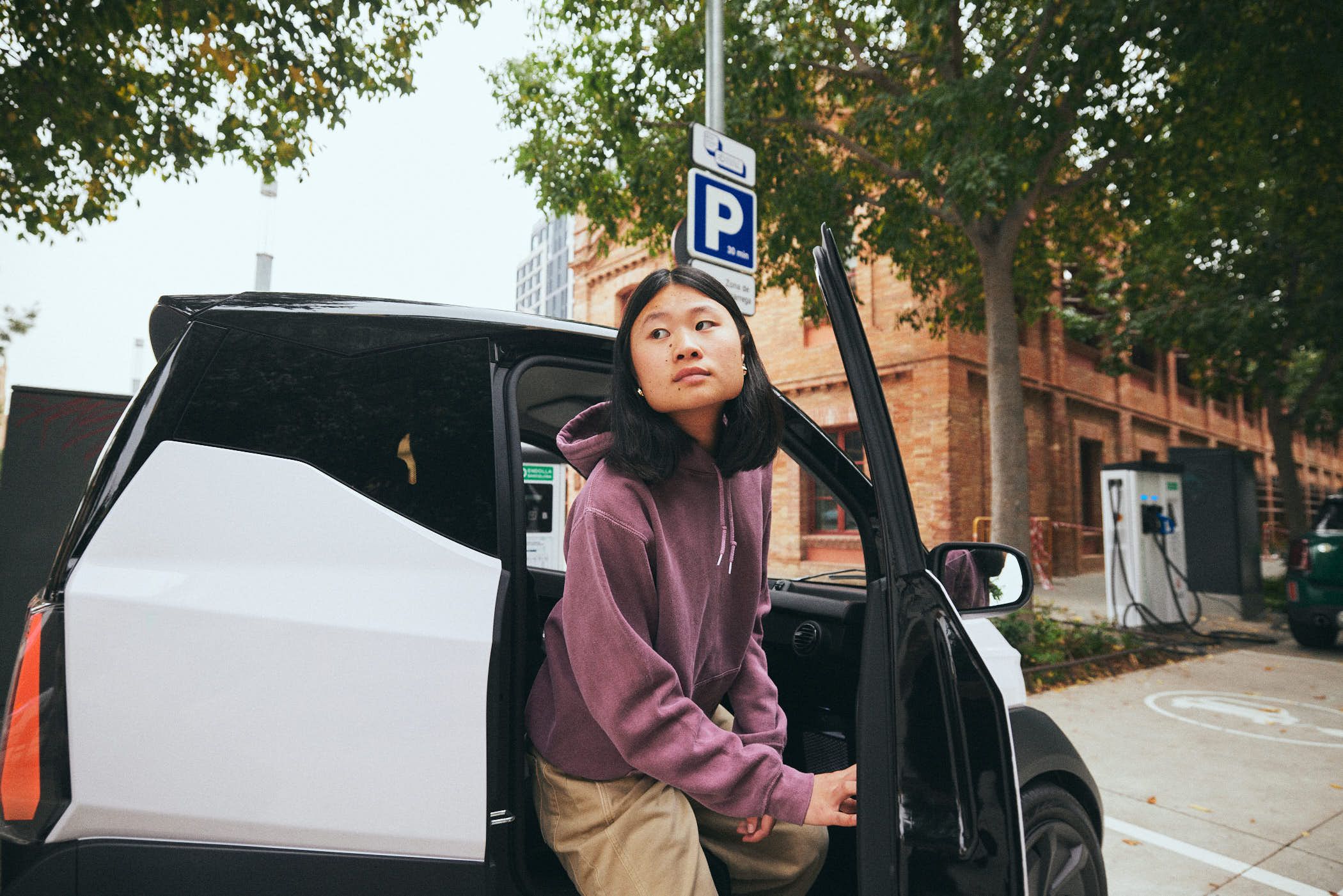 A young Asian girl with a neutral expression is getting into the passenger side of a white car with the door open. She is wearing a purple hoodie and khaki pants, and the car is.