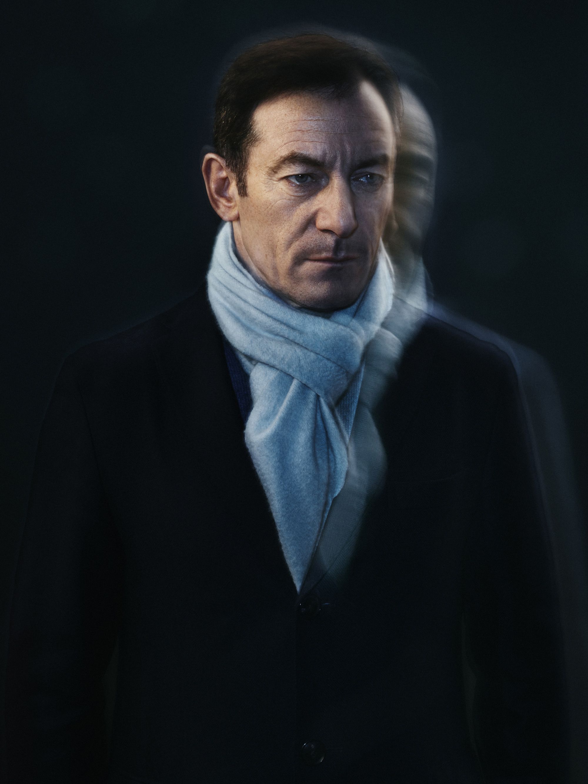 A man in a black suit and white scarf, looking directly at the camera with a serious expression.