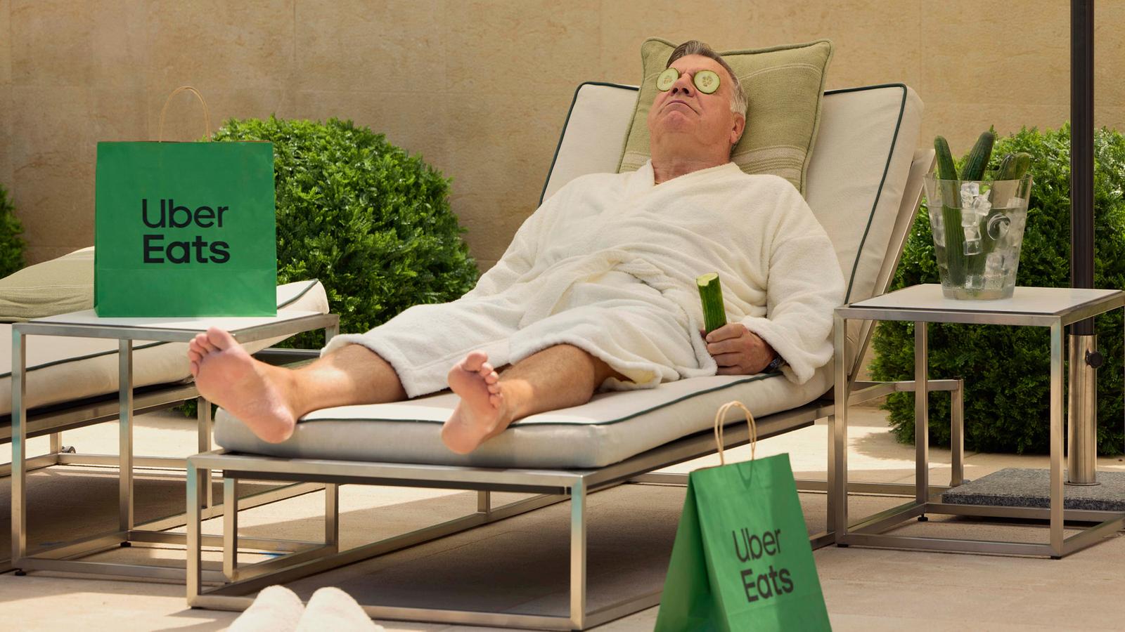 A man in a white bathrobe, lounging on a couch, with a food delivery bag from Uber Eats on the table next to him, and a cucumber slice on his eye, in a luxurious outdoor setting.
