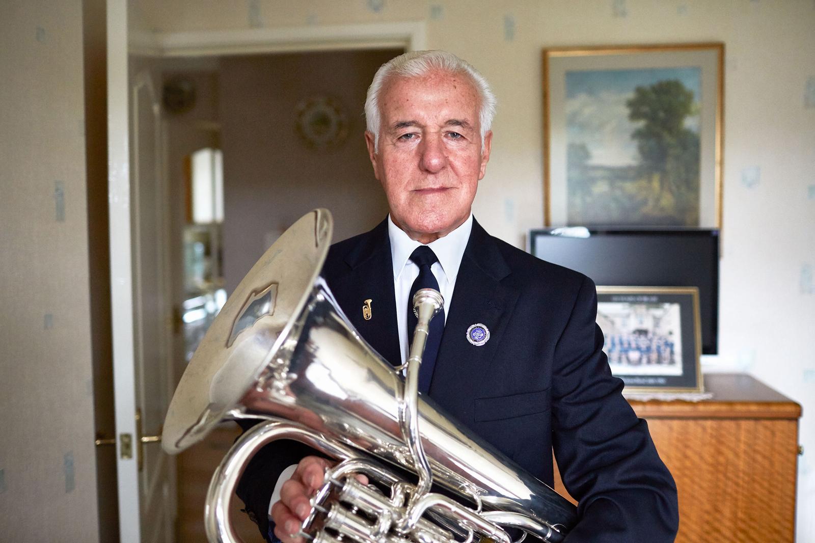 A man in a suit and tie holding a silver trumpet, with a background of a room with a painting and a bookshelf.