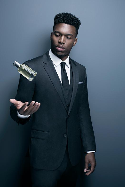 A man in a black suit, white shirt, and black tie, holding a bottle of champagne in his hand, with a serious expression on his face.