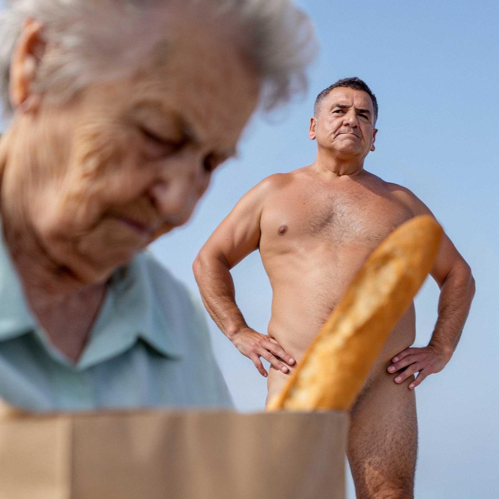 The image shows an older woman in the foreground wearing a light green shirt, looking down into a grocery bag. In the background, a naked man stands with his hands on his hips, looking off into the distance. A baguette sticks out prominently from the grocery bag in the foreground, partially obscuring the man's lower body.