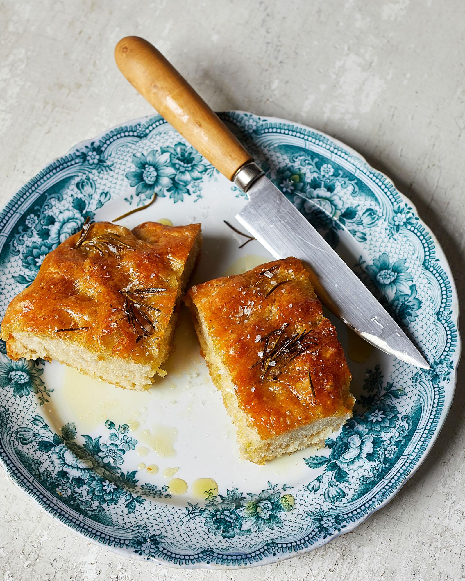 A slice of golden-brown, crispy focaccia bread, topped with olive oil and rosemary, on a white plate with a blue floral pattern.