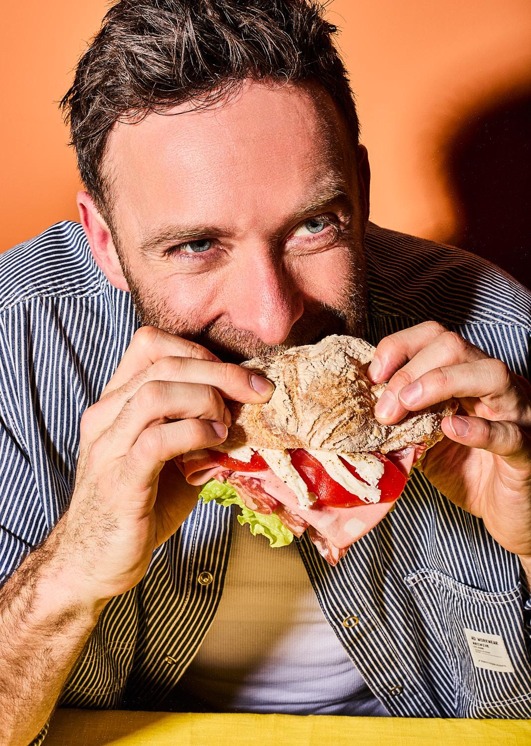 The image shows a close-up of a man in a blue striped shirt taking a large bite of a sandwich. He is looking off-camera with an intense, satisfied expression, holding the crusty bread filled with tomatoes and mozzarella with both hands.