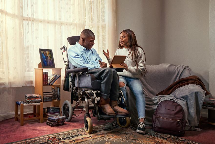A man in a wheelchair, wearing a blue shirt and jeans, sitting in a room with a woman holding a tablet, in a home setting.