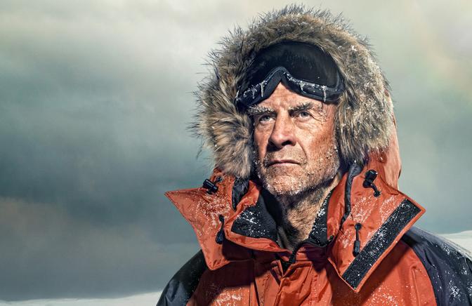 A man in a thick, orange and black parka with a fur-lined hood, snow-covered and ruggedly weathered, standing in a snowy landscape.