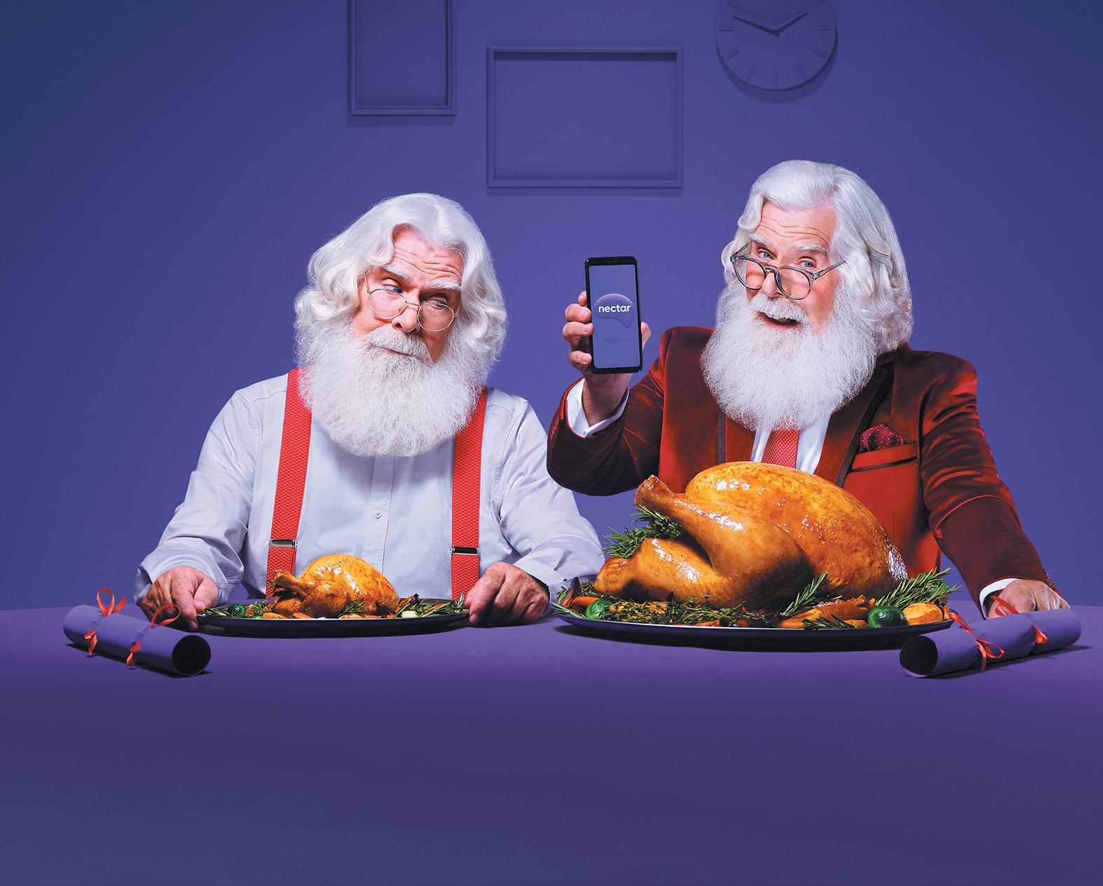 Two Santa Clauses, one in a white shirt and red suspenders, and the other in a red suit, sitting at a table with a large turkey and a smartphone displaying the Nectar logo.