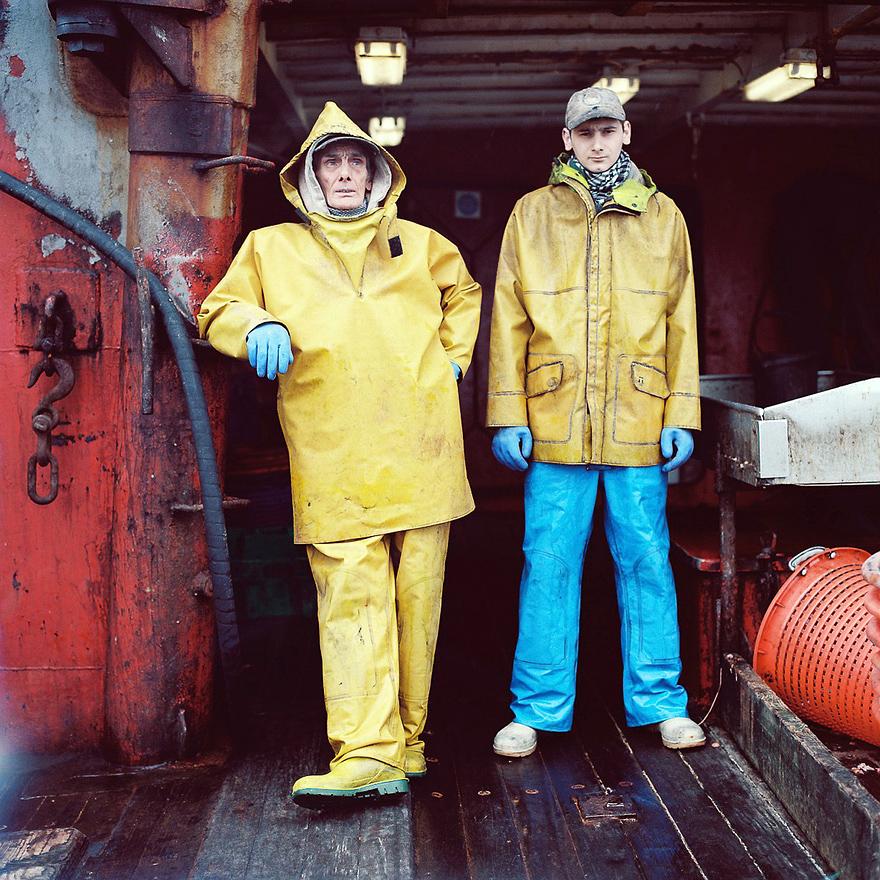 Two men in yellow raincoats and blue rubber gloves stand on a boat.