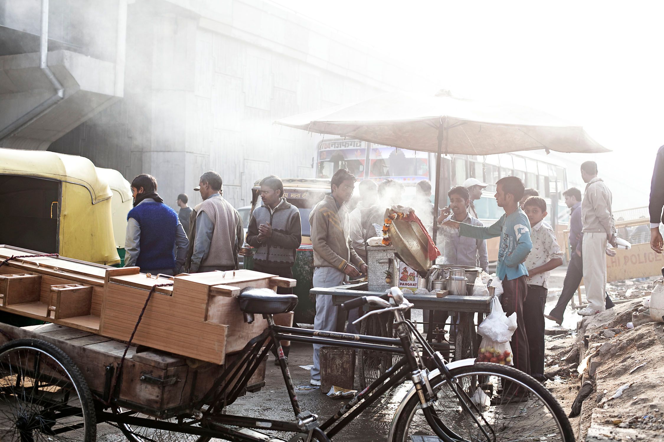 A bustling street scene in India, with a food cart and a crowd of people in the background, and a yellow auto-rickshaw and a black bicycle in the foreground.